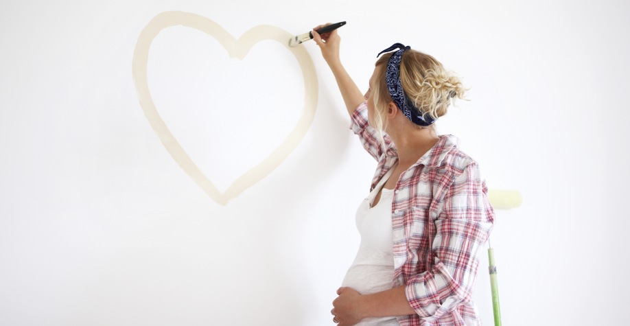 Eine schwangere Frau malt mit einem Pinsel ein Herz an eine weiße Wand.
