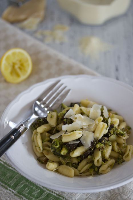 Lustig leicht eingedrehte Nudeln mit grünem Pesto in einem feinen Teller. Im Hintergrund liegt eine Zitrone und Grieß.