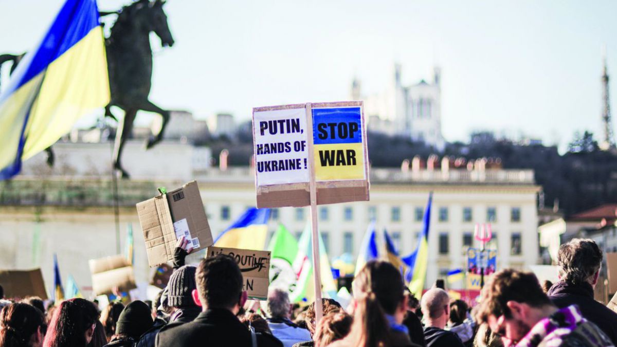 Menschen demonstieren mit blau-gelben Fahnen und Transparenten gegen den russischen Angriff auf die Ukraine. Auf einem Schild steht: 