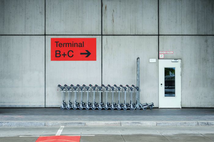 Gepäcktrolleys vor dem Flughafengebäude. Ein oranges Schild an der Betonwand trägt Aufschrift mit Richtungspfeil:  Terminal B+C.