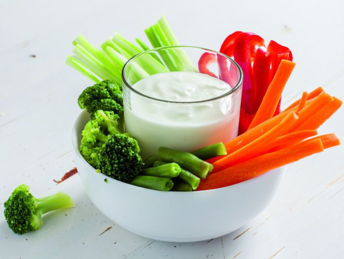 Ein Glas voll Joghurt-Topfen-Dip in einer Schale mit Gemüsesticks: Selleriestangen, Karotten, Fisolen, Rote Paprika, Brokkoli.