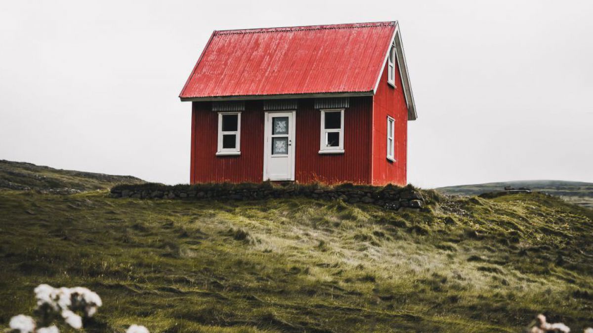 Ein kleines, rotgestrichenes Holzhaus mit weißen Fenstern und rotem Dach steht einsam in einer hügeligen, grasbewachsenen Landschaft. Im Bildvordergrund blüht Schafgarbe.