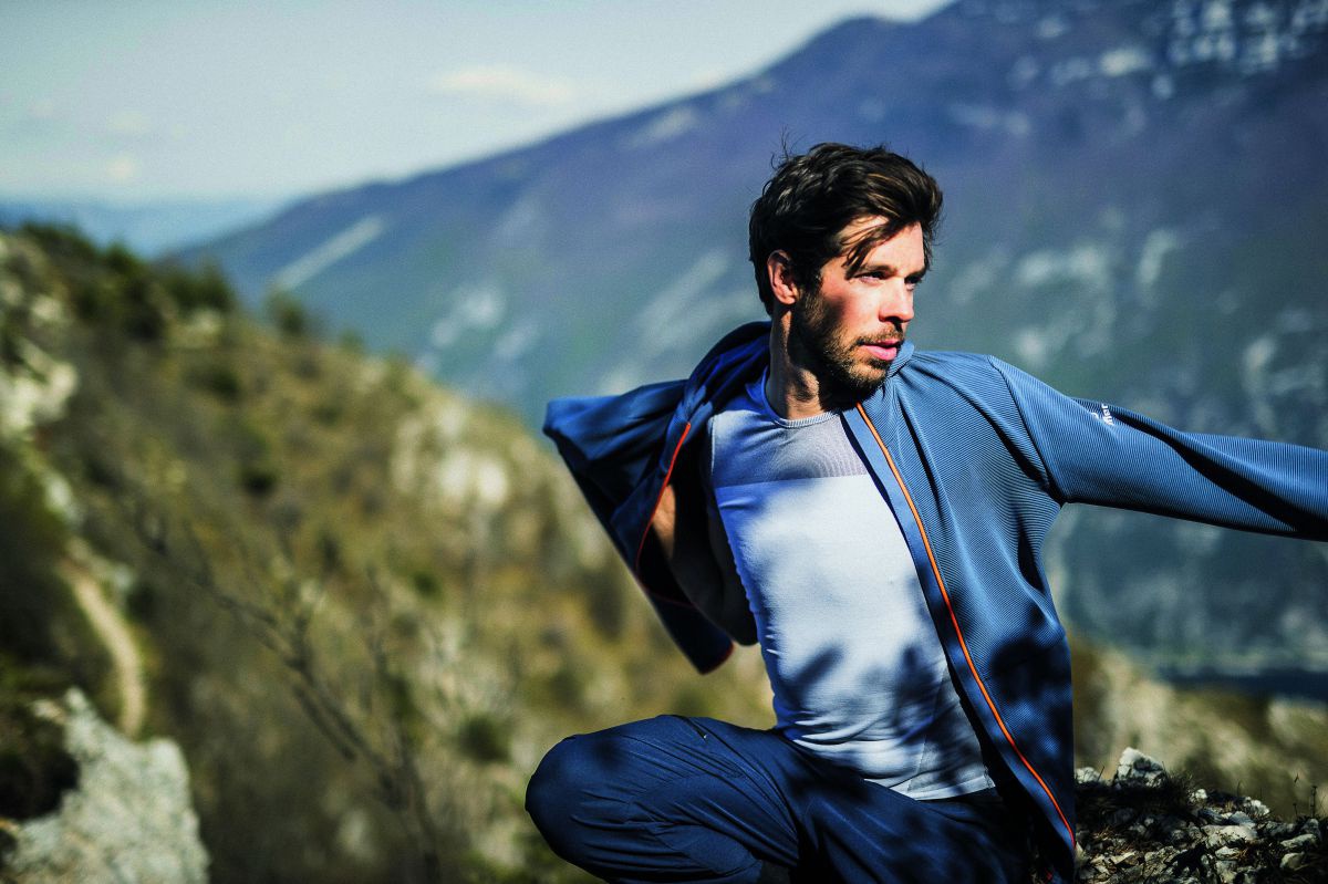 Ein junger Mann zieht sich gerade seine Sportjacke an. Er hält sich im Gebirge auf, im Hintergrund sieht man bewaldete Berghänge und felsige Gebirgsrücken.