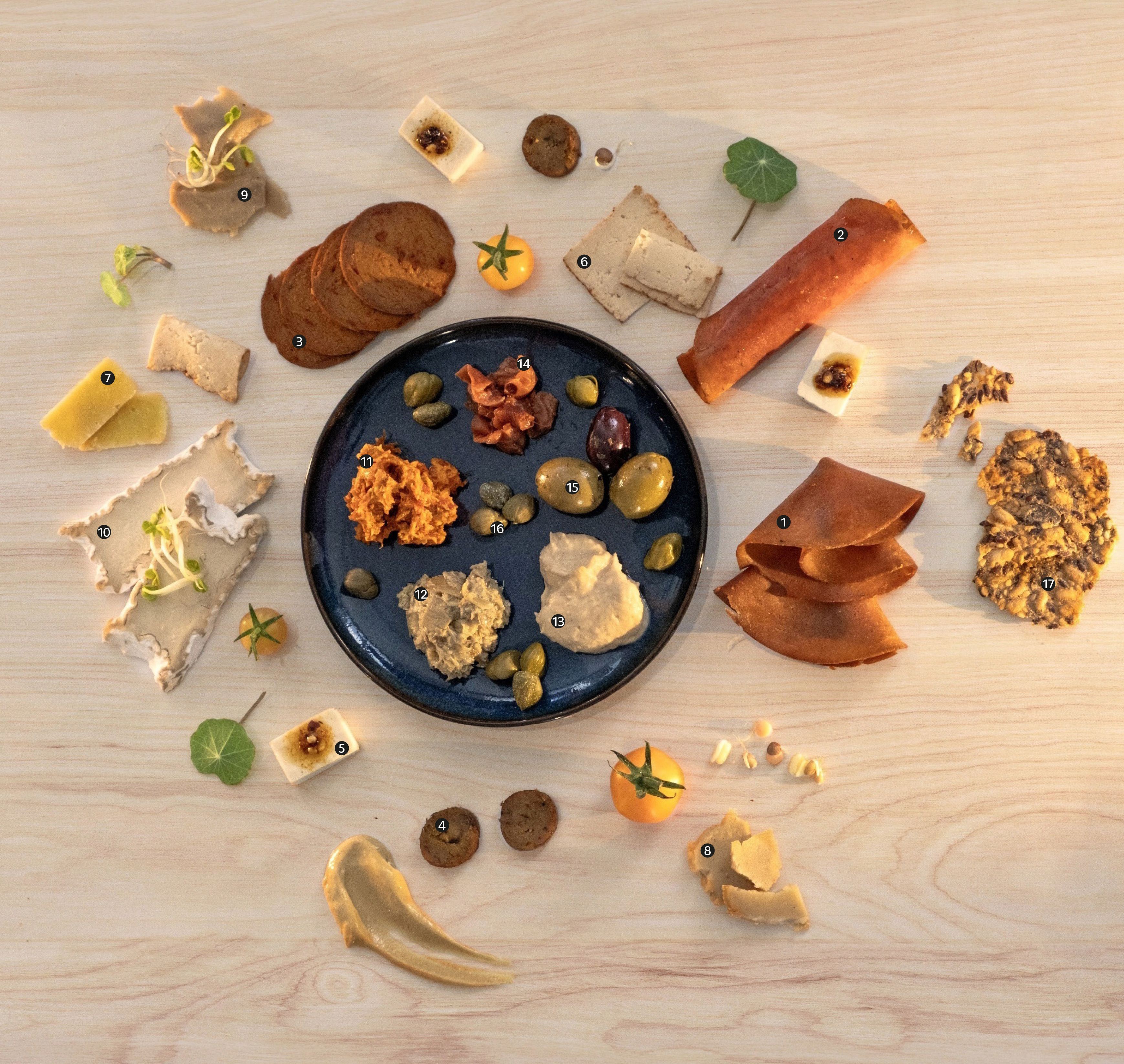 Auf einem hellen Holztisch sind verschiedene kleine Portionen von Aufstrichen, Wurstscheiben, Käse, Früchten, Sprossen und Kräcker dekorativ rund um einen blauen Teller mit Kapern, Oliven und Dips angerichtet.