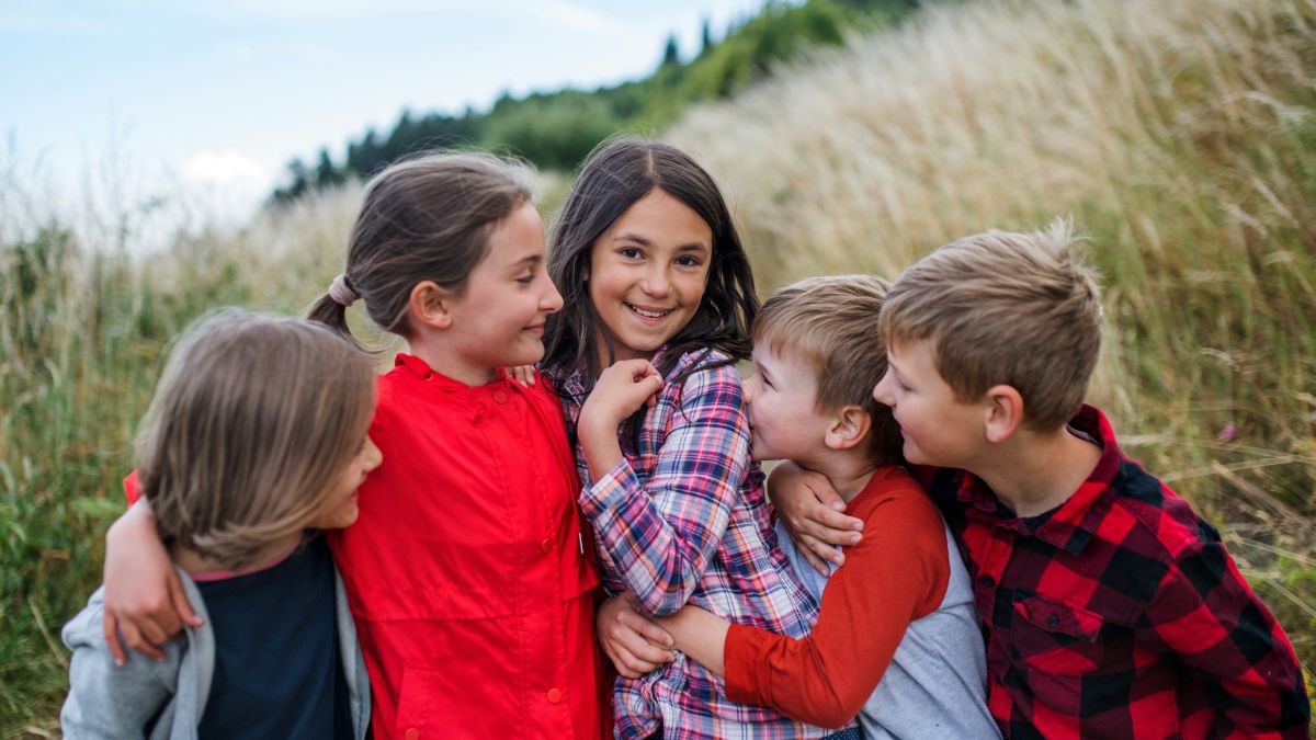 Vier Geschwister oder Freund*innen im Schulalter stehen dicht aneinander gedrängt in einer Gruppenumarmung im Freien, ringsherum sieht man hohes, blühendes Gras und etwas entfernt einen Nadelwald am Horizont. Die Kinder wirken glücklich aufgeregt.