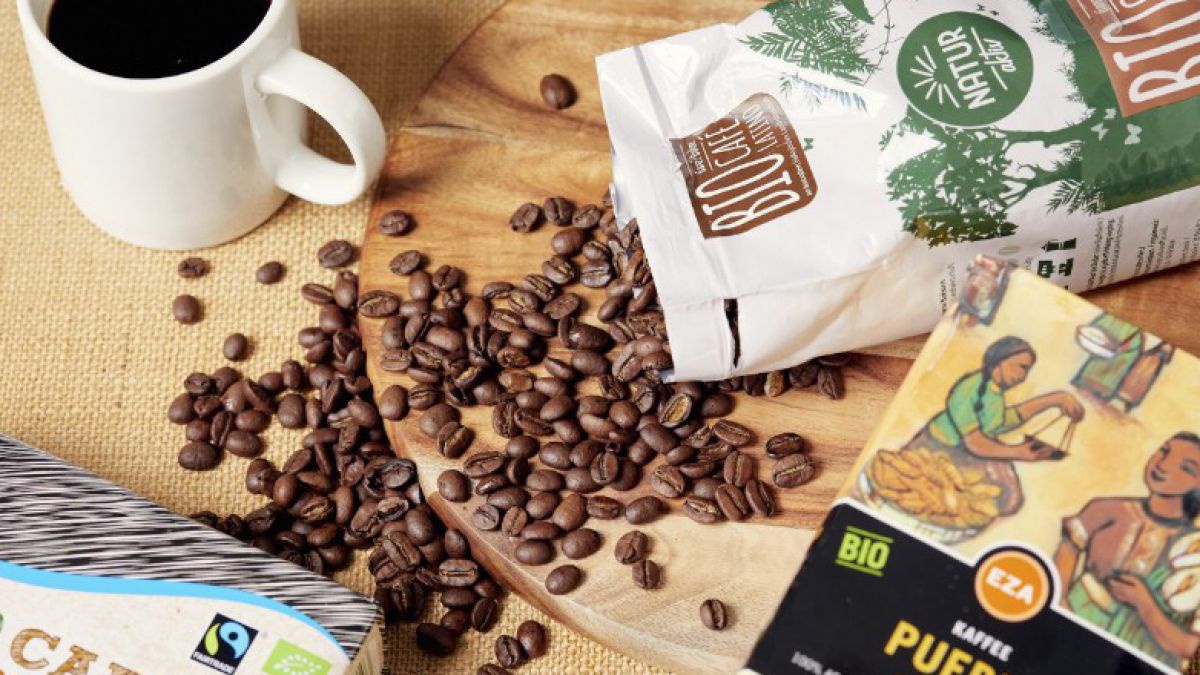 Aus einer offenen Kaffepackung ergießen sich Bohnen auf den Tisch. Eine Kaffeetasse und andere Kaffeepackungen sind rundherum zu sehen.