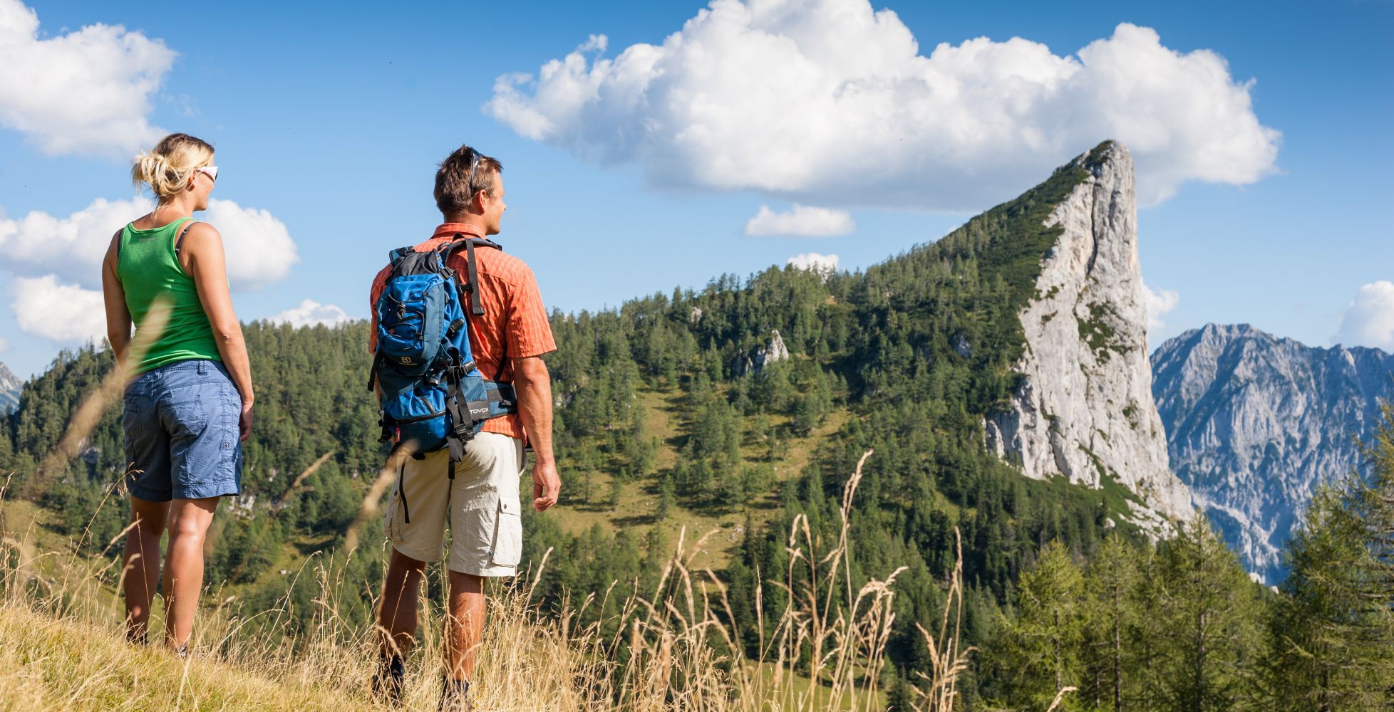 Ein Mann und eine Frau stehen auf einer Wiese. Sie blicken auf tolles Bergpanorama.