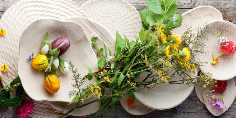 Auf beigen Tellern in verschiedenen Formen liegen bunte Gemüse, Kräuter und Blüten.