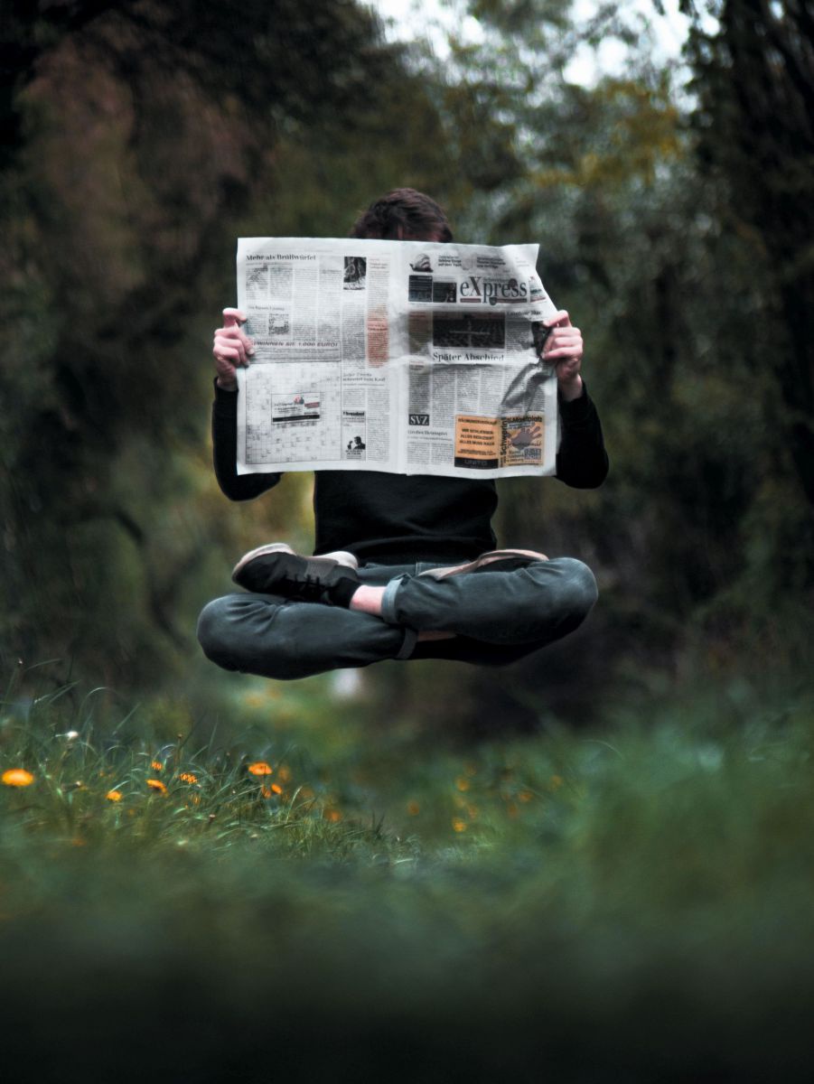 Ein junger Mann schwebt im Schneidersitz über dem Gras. Er hält eine Zeitung lesend vor dem Körper.