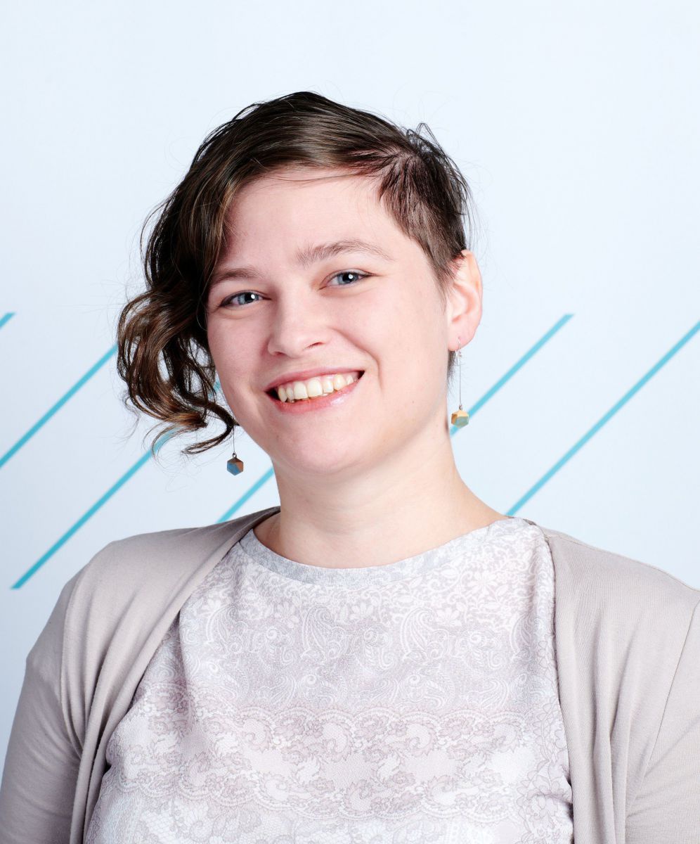 Michaela Reisinger ist Mitte 30, dunkelbraune Haare lockig auf einer Seite kinnlang, auf der anderen kurz, blaue Augen, trägt beige Kleidung, lacht in die Kamera.