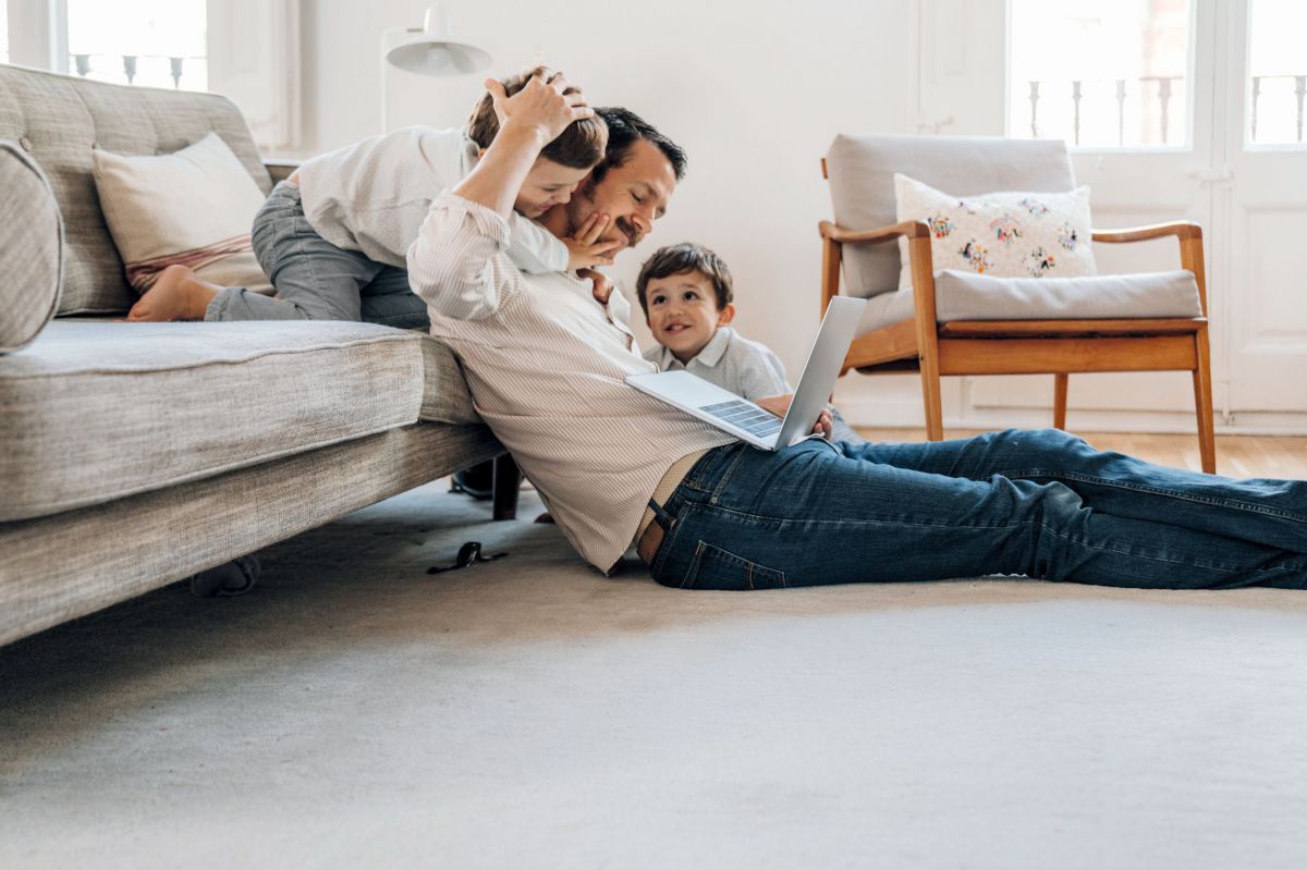 Ein Vater sitzt am Boden vor der Couch, den Laptop am Bauch. Ein Kind sitzt neben ihm, eines schaut ihm über die Schulter. Eine fröhliche Familienszene.