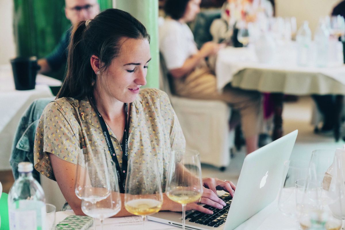 Juliane Fischer sitzt an einem gedeckten Tisch und schreibt auf ihrem Laptop mit. Vor ihr stehen vier Weingläser.