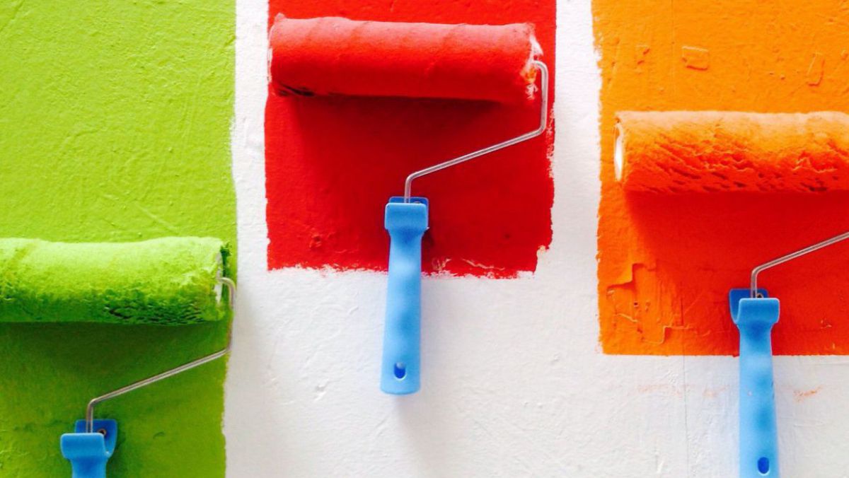Mit drei Farbrollern wird grüne, rote und orange Farbe auf eine weiße Wand aufgetragen.