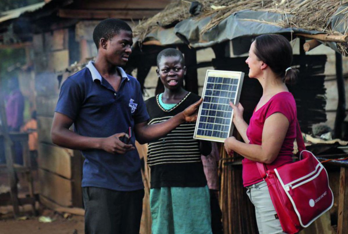 Zwei jüngere schwaze Menschen und eine weiße Frau unterhalten sich über ein zeitungsgroßes Solarpanel. Im Hintergrund ist eine rurale Hütte zu sehen.