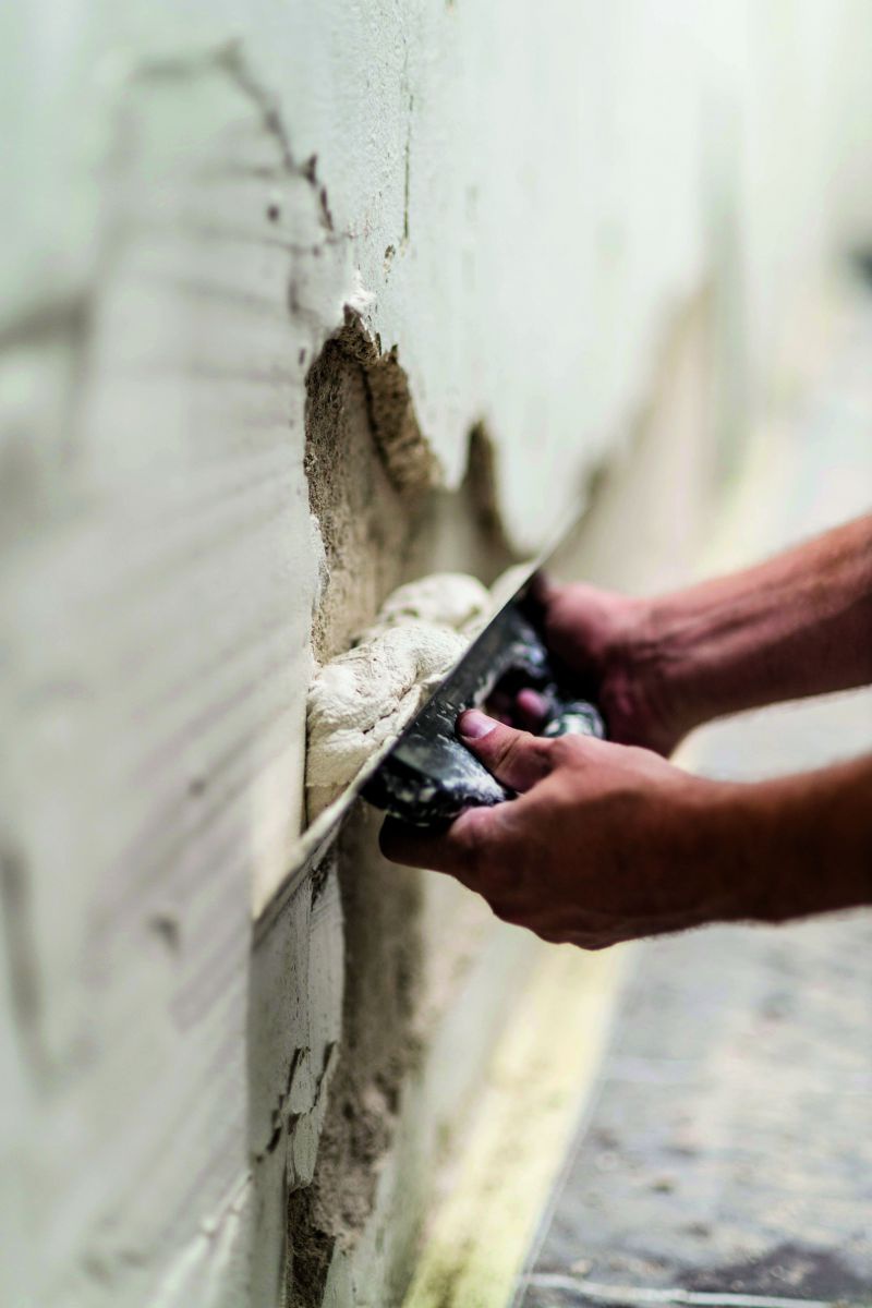 Zwei Hände füllen mit einem großen Spachtel fehlenden Putz an einer Wand auf.