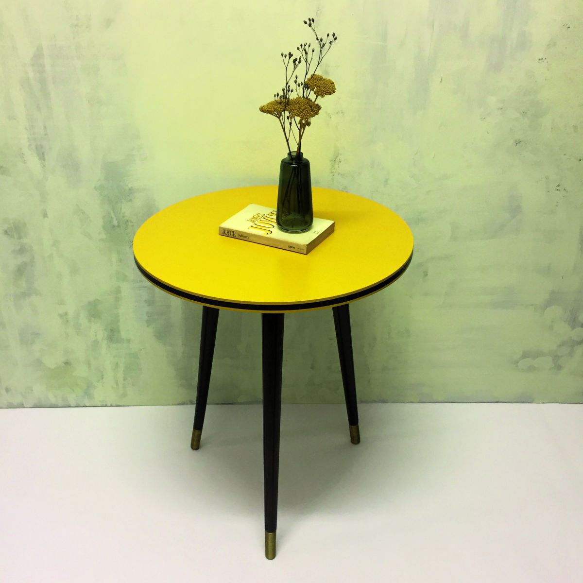 Ein runder dreibeiniger Vintagetisch hat einen neuen senfgelben Anstrich bekommen. Auf dem Tisch liegt ein Buch, darauf steht eine Vase mit Trockenblumen.