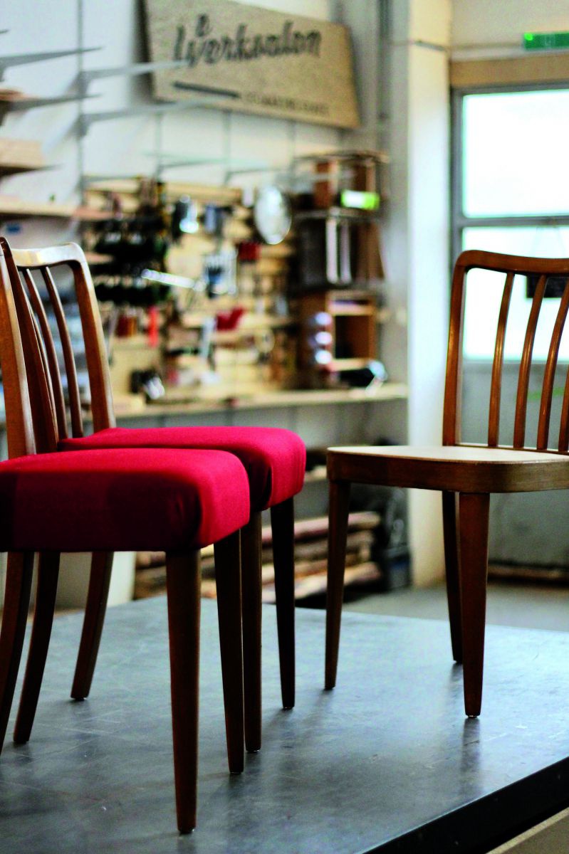 Drei Sessel mit geschwungenen Rückenlehnen - zwei mit rotem Bezug, einer ganz aus Holz - stehen in einer Werkstatt.