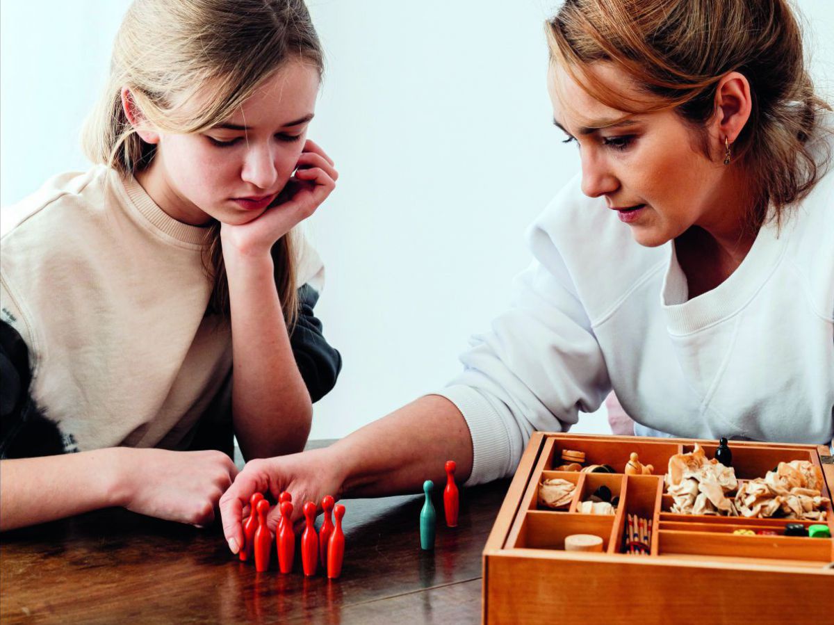 Eine Frau und eine Jugendliche stellen aus einer Box Spielfiguren auf einem Tisch auf. Sie wirken nachdenklich.