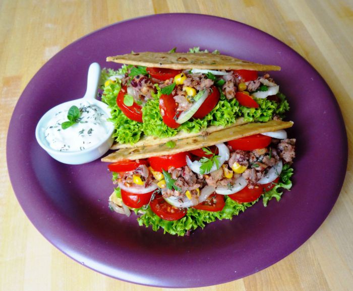 Zwei mit Salat, Tomaten, Zwiebeln, Mais und Bohnen gefüllte Tacos auf einem lila Teller. Daneben ein Schälchen mit Kräutersauce.