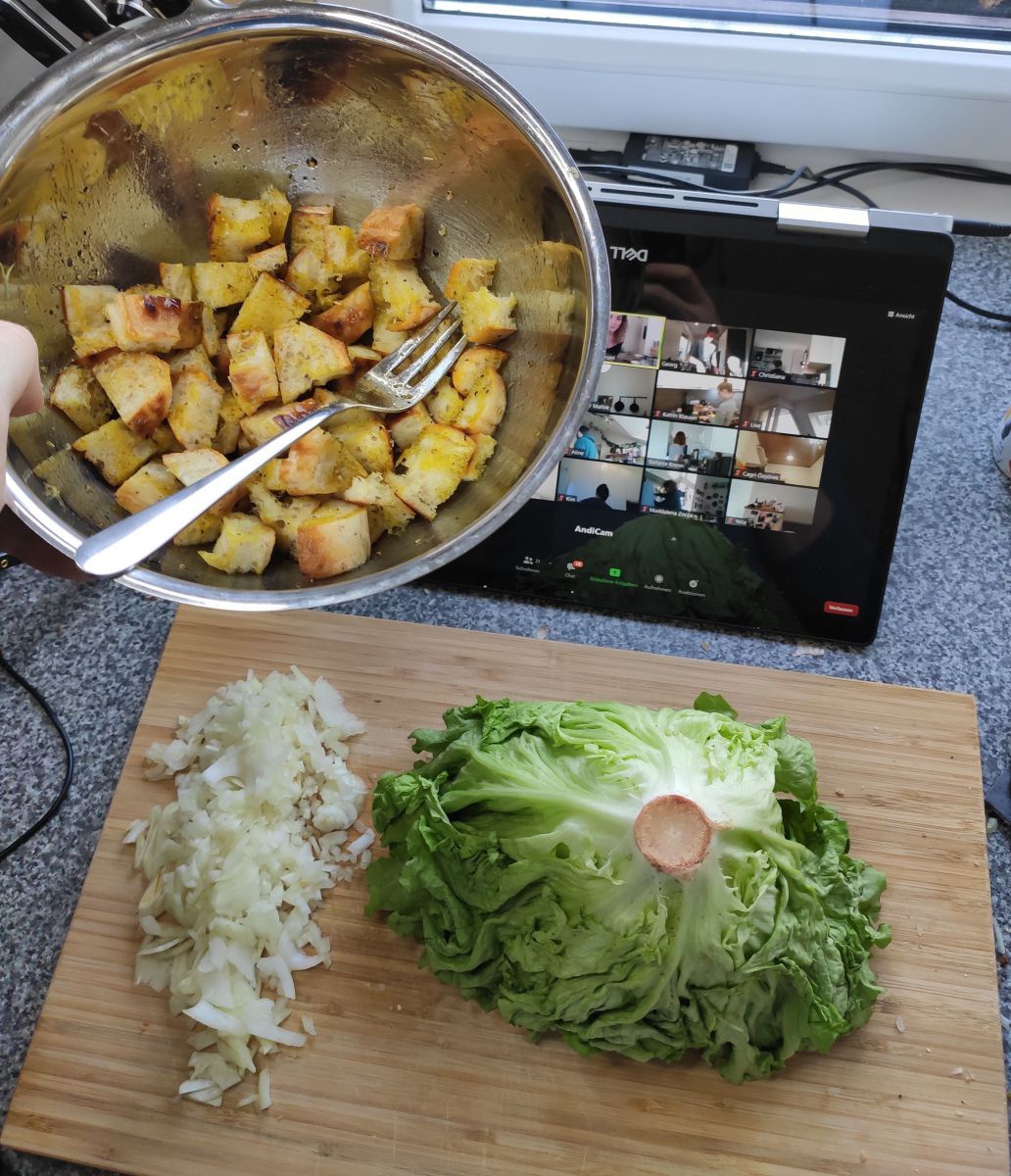 Jemand hält eine Schüssel mit Croutons vor die Kamera. Dahinter ist ein Shneidbrett mit einem Salatkopf und geschnittenen Zwiebel sowie ein Tablet mit einer laufenden Videokonferenz zu sehen.