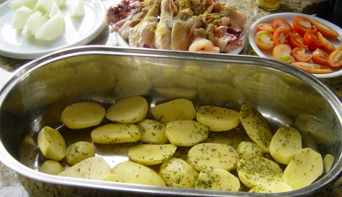 Eine ovale Ofenform, mit Kartoffel ausgelegt. Dahinter stehen drei Teller mit Zwiebeln, Hühnchenteilen und Tomaten.