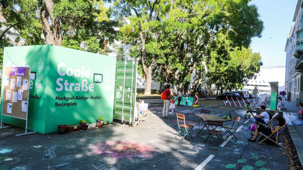 Ein Container, Tische und Sessel auf einer abgesperrten Straße. Der Asphalt ist mit Kreide bunt bemalt. Auf einer Straßenseite sind Bäume.