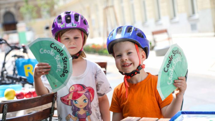 Zwei Kinder mit Fahrradhelmen haben grüne Papierfächer in der Hand, auf denen 