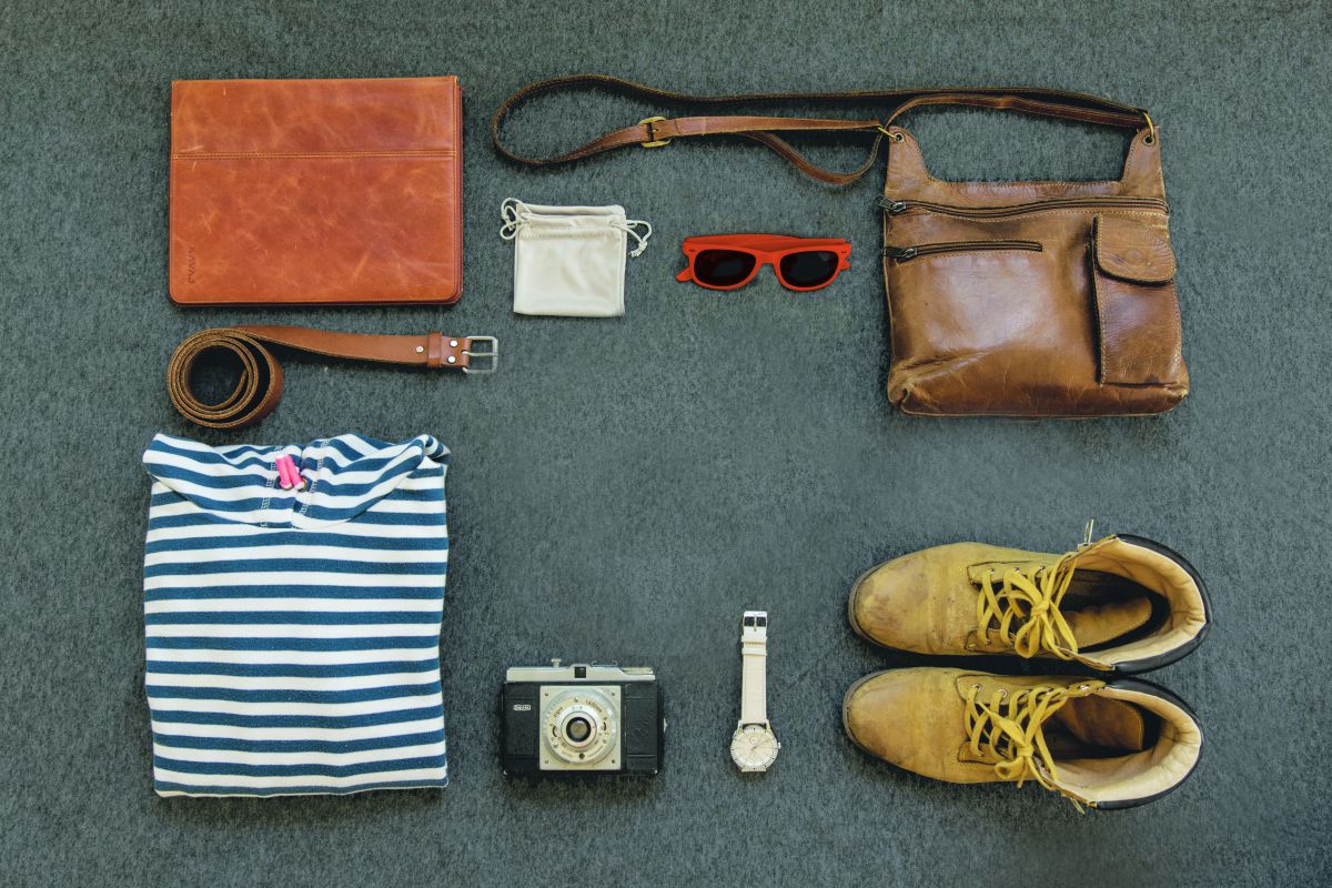 Auf einer Oberfläche sind Sachen aufgelegt: ein Gürtel, eine Sonnenbrille, Schuhe, eine Tasche, eine Armbanduhr, ein Tablet, ein Beutel und eine Kamera.