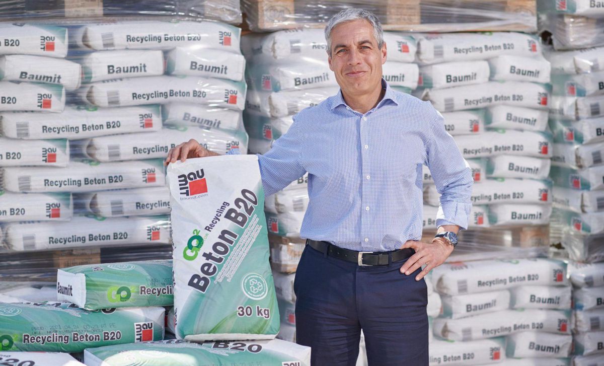 Ein Mann steht vor Paletten mit Betonsäcken und hält einen aufgestellt gegen seinen Arm gelehnt.