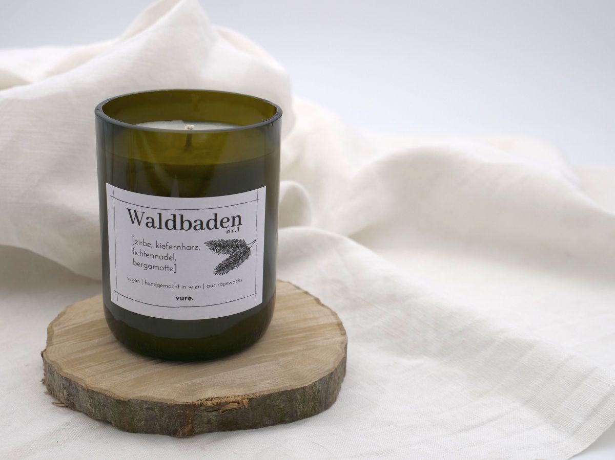 Eine Kerze mit weißem Wachs in einem dunkelbraunen Glas steht auf einem Holzuntersetzer. Auf der Kerze klebt ein Etikett 