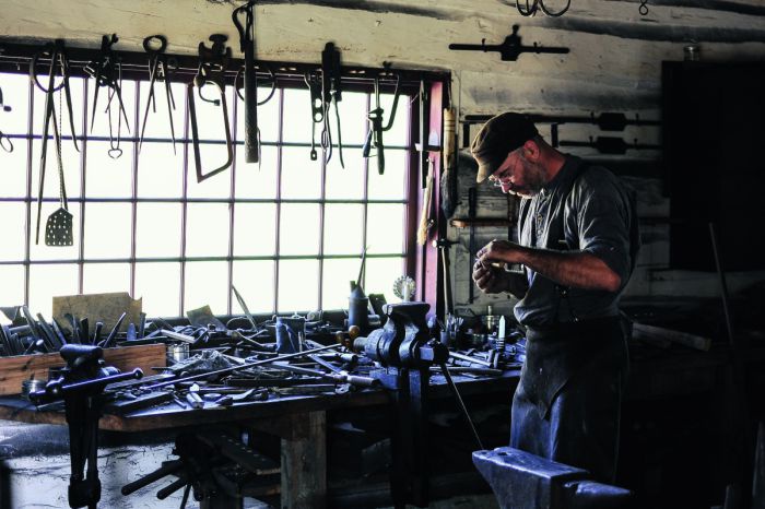Ein Mann steht in einer Werkstatt. An der Decke hängt Werkzeug, der Arbeitstisch ist voll geräumt. Der Mann bearbeitet etwas in seinen Händen.