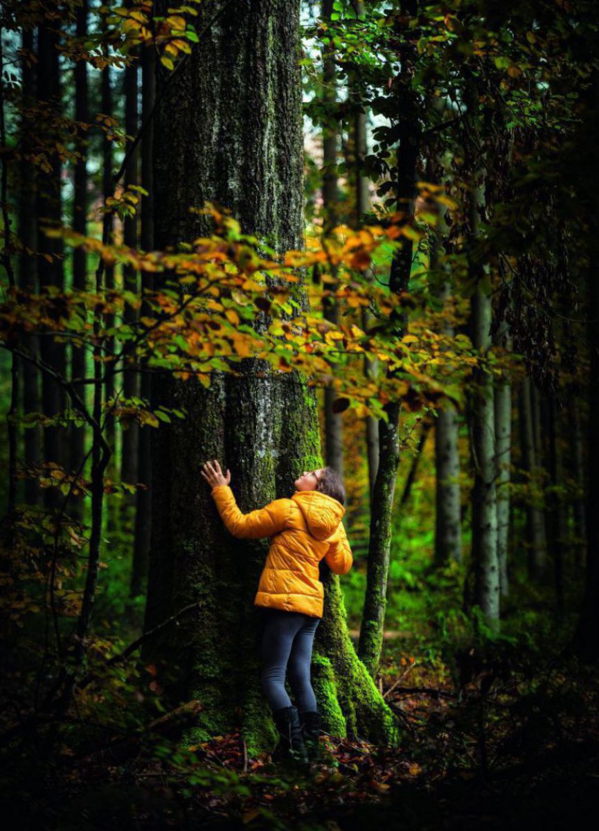 Eine Jugendliche in gelber Daunenjacke umarmt einen Baum.