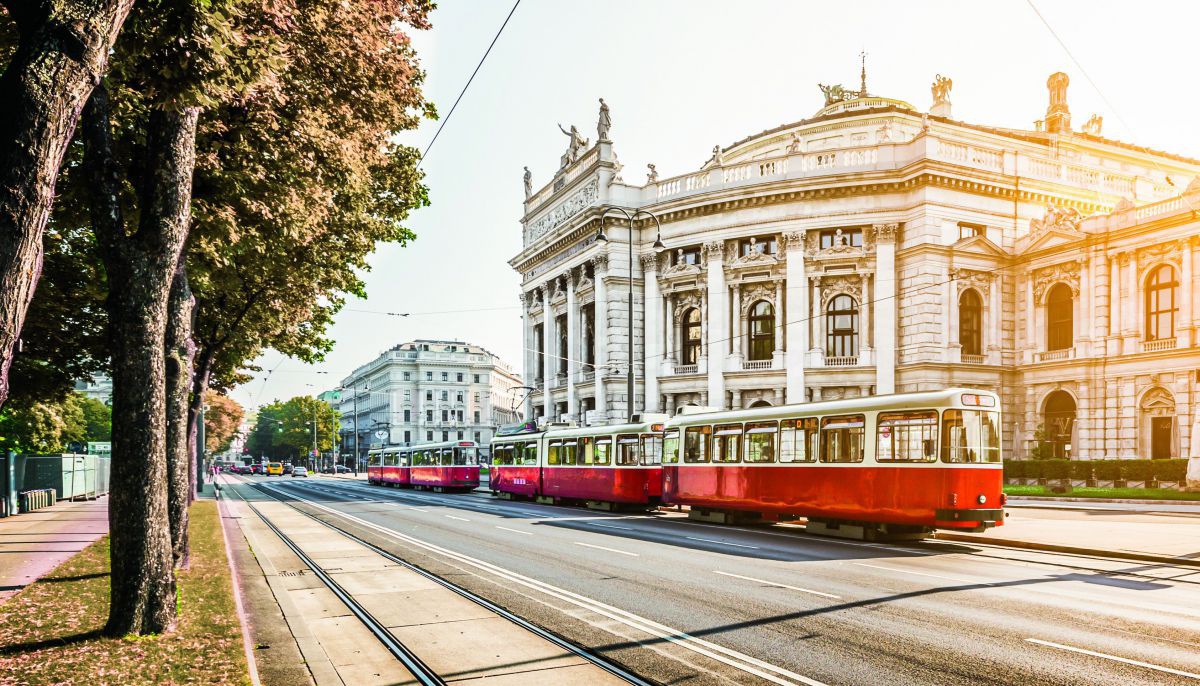 Ein Bild vom Wiener Ring. Rechts das Volkstheater links einige Bäume. Auf den Fahrspuren Autos und eine Straßenbahn.