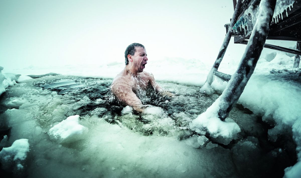 Ein Mann taucht aus einem See. Die Leiter ist gefroren, im Wasser schwimmt Eis.