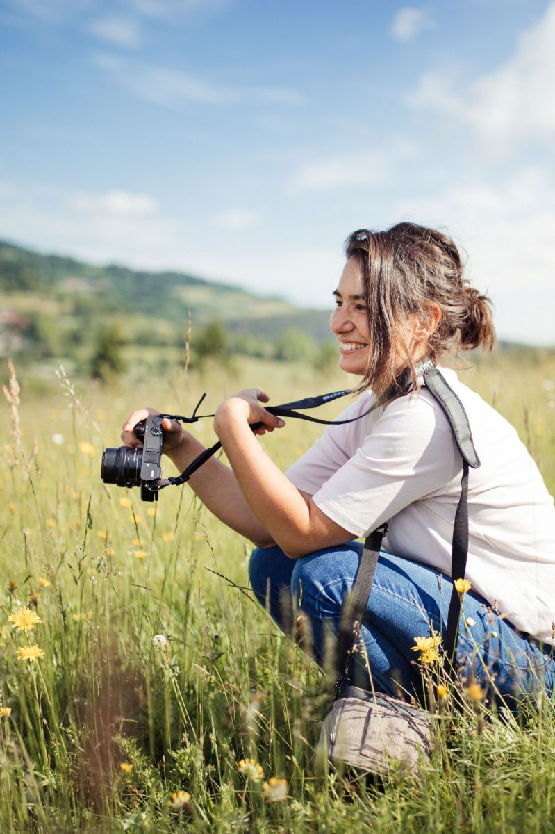 Mathilde Stallegger hockt in einer Blumenwiese, in der Hand ein Fotoapperat. Die Sonne scheint, sie lacht.