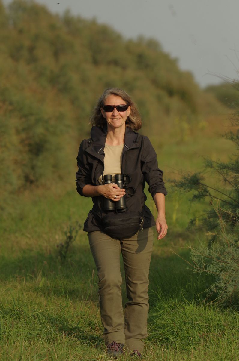 Birgit Forthuber geht im Foto auf die Kamera zu. Sie trägt eine Sonnenbrille und ein Fernglas um den Hals. Im Hintergrund Natur. 