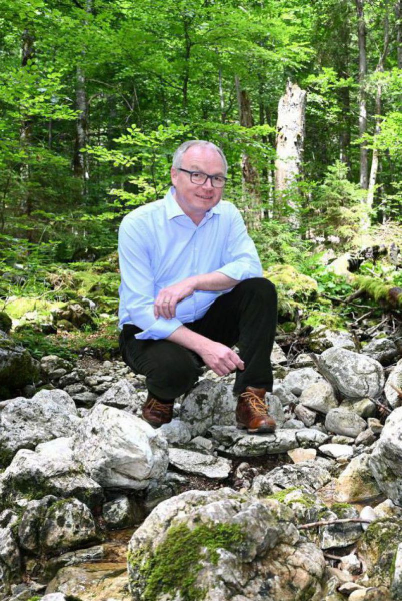 Stephan Pernkopf kniet in einem Wald an einem Bächlein. Er trägt eine Brille und ein hellblaues Hemd.