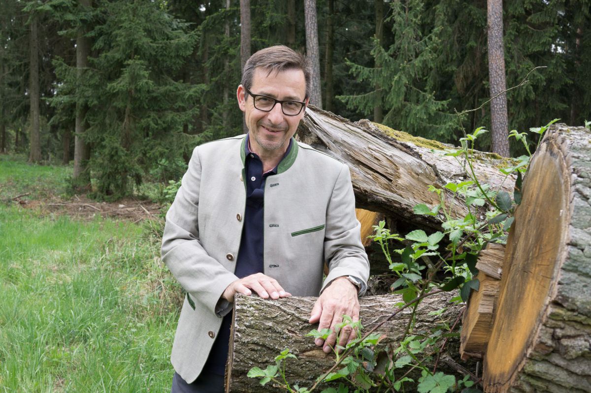 Herbert Schmidt steht am Waldrand neben einem Holzstoß. Er trägt Brille und ein Trachtensakko.