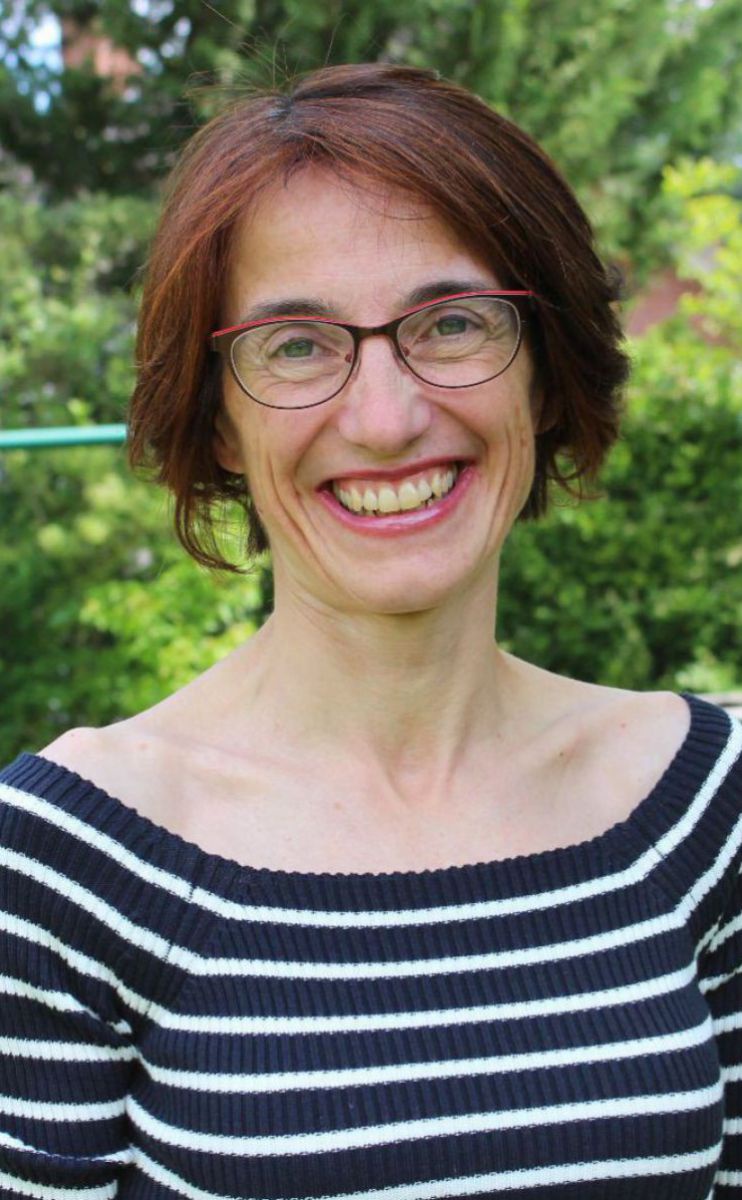Julia Kelemen-Finan auf einem Portraitfoto in der Natur. Sie hat rötliche Haare, trägt eine Brille und lacht in die Kamera.