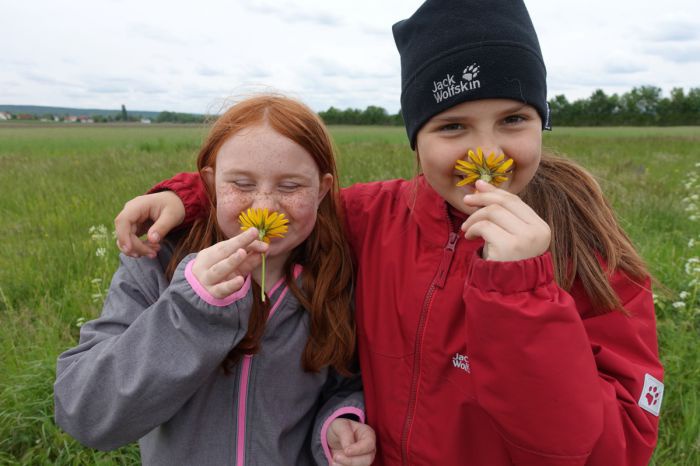Zwei Mädchen schnuppern an gelben Blüten. Sie lachen, das eine legt den Arm um die Schultern des anderen.