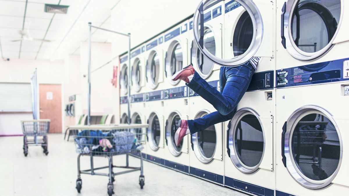 Eine lange Reihe mit Waschmaschinen in einem Waschsalon. Eine Person hängt zur Hälfte in einer Trommel der oberen Reihe. Nur ihre Beine und Füße schauen heraus.