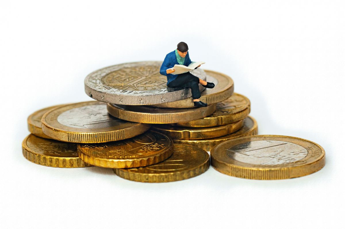 Eine kleine zeitungslesende Figur sitzt mit überschlagenen Beinen auf einem Haufen Euro-Münzen.