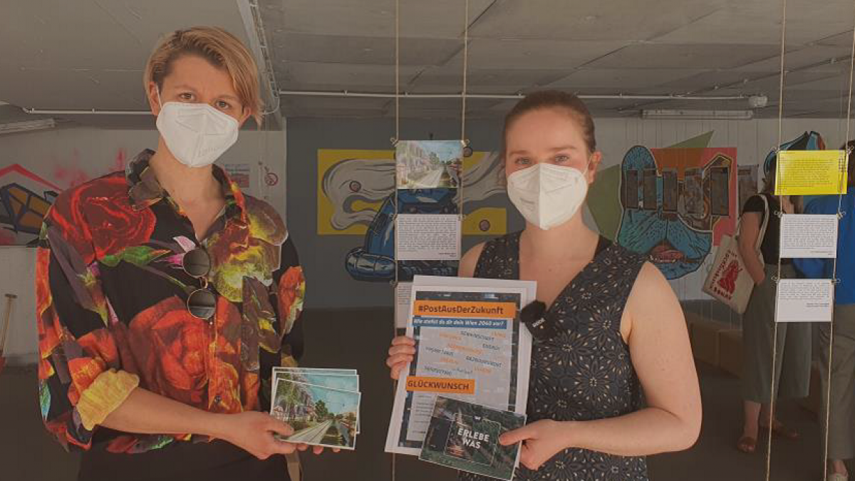 Preisverleihung von klimaaktiv: Zwei Frauen stehen nebeneinander und halten Bilder und ein Zertifikat in die Kamera. Im Hintergrund ist eine Ausstellung zu sehen.