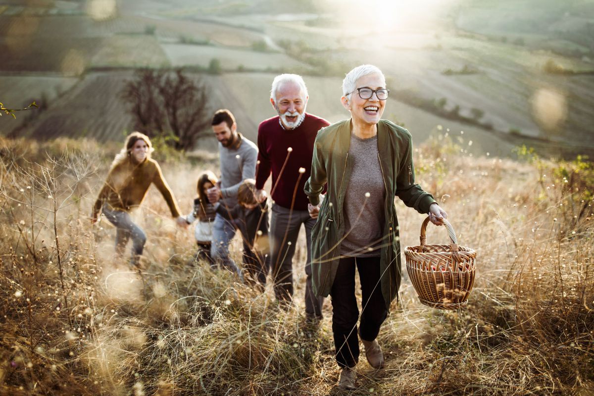 Eine Familie geht hintereinander über eine Wiese mit hohem Gras. Sie halten einander an den Händen. Die Großeltern mit weißen Haaren führen, dahinter jeweils die Enkelkinder und erwachsenen Kinder.