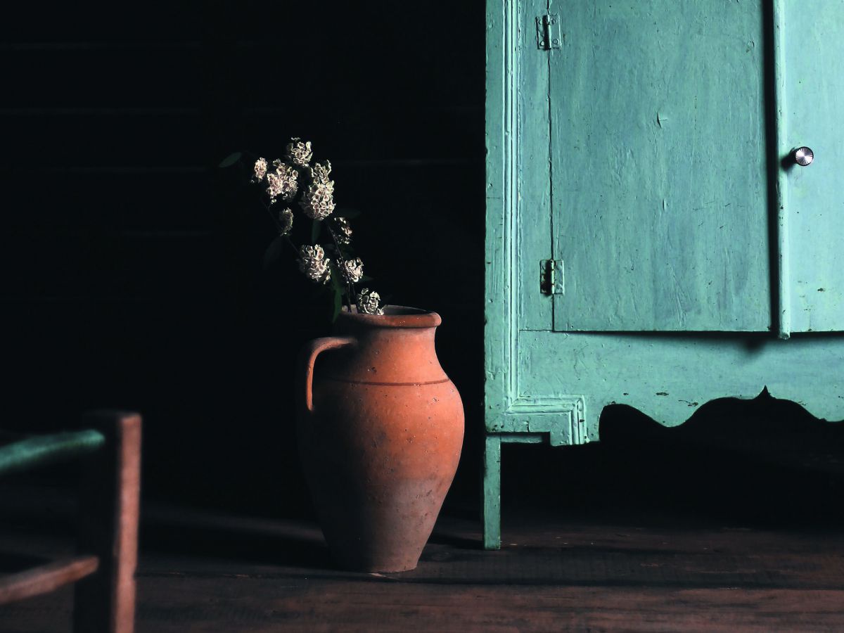 Ein Tonkrug mit verblühten weißen Blumen steht nehmen einer Holzanrichte. Starke Kontraste, dunkler Hintergrund.