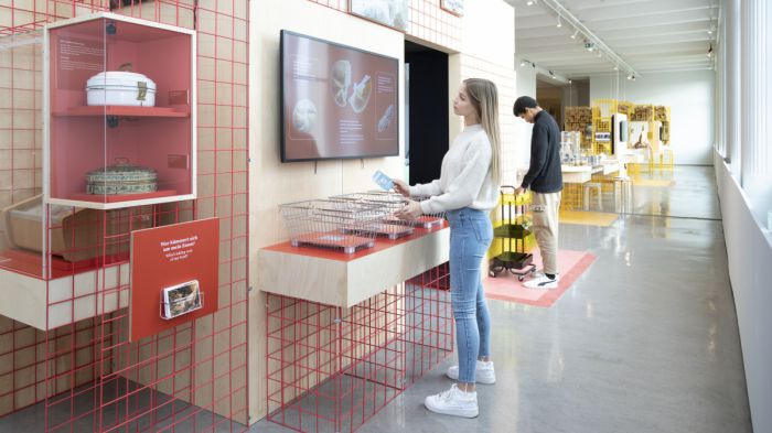 Im Ausstellungsraum ist eine rote Gitterwand und ein Raum aufgebaut. Auf der Außenseite ist ein Screen mit drei Interaktionspunkten aufgebaut.