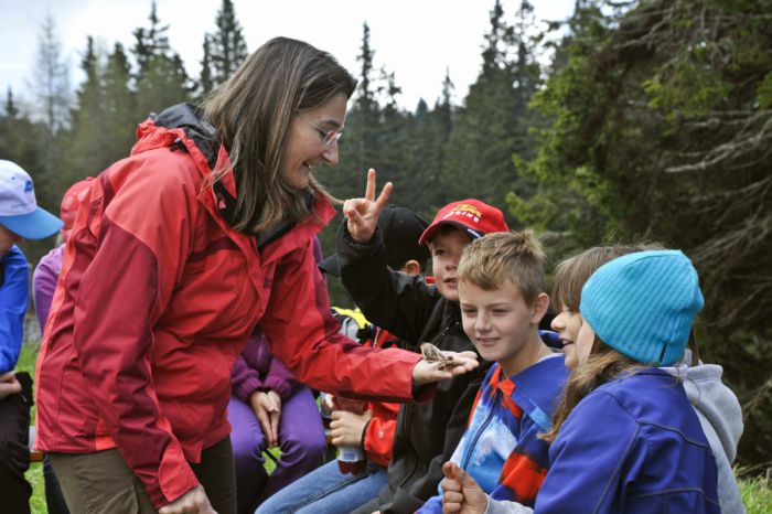 Eine Frau hält einen Falter auf ihrer flachen Hand um ihn einer Gruppe von Schulkindern zu zeigen, im Hintergrund ein Nadelwald. 