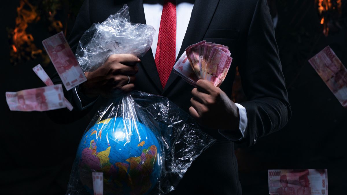 Ein Mensch mit Anzug und Kravatte. Er hat einen Starbucks-Sack über den Kopf gestülpt und hält in der einen Hand einen durchsichtigen Plastiksack mit einer Weltkugel, in der anderen Geld. Im Hintergrund brennt es.