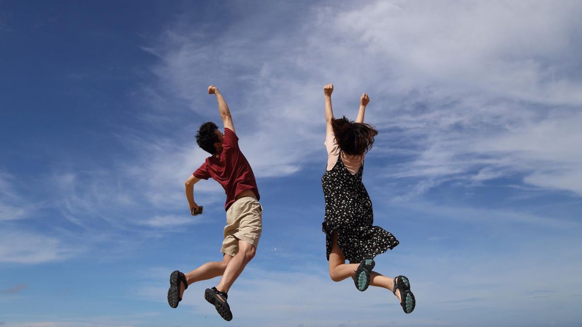 Zwei Menschen springen mit in die Höhe gestreckten Armen in die Luft, im Hintergrund ein sanft bewölkter blauer Himmel.