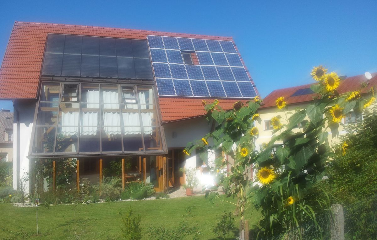 Man sieht ein Haus mit einem Wintergarten, einer thermischen und einer PV-Anlage. Im Vordergrund eine Sonnenblume